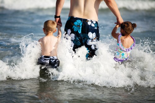 A man and two children wade into the ocean, splashing water as they enjoy a sunny beach day.