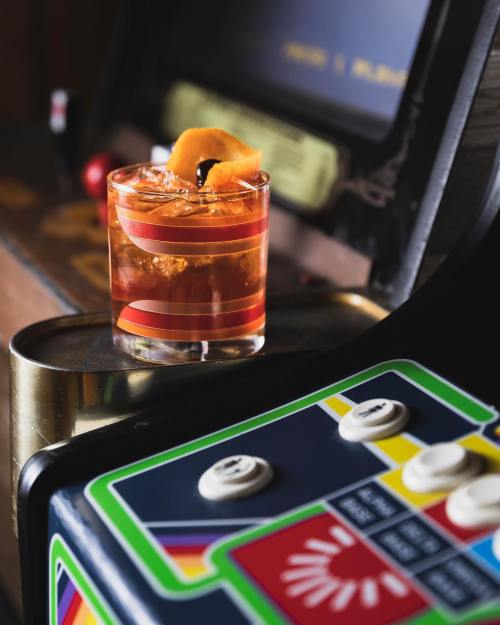 A cocktail with an orange twist sits on a bar top next to a vintage arcade game.