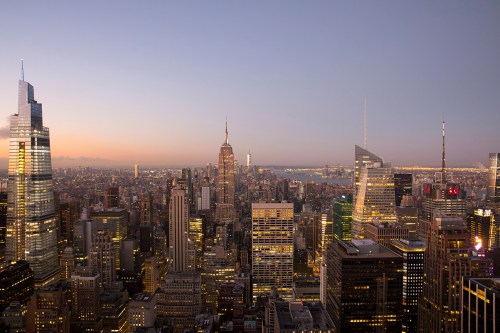 A panoramic view of a city skyline at dusk, featuring tall buildings and the Empire State Building illuminated.
