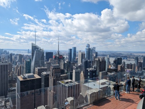 A panoramic view of a city skyline with skyscrapers and a cloudy sky, featuring people on a observation deck.