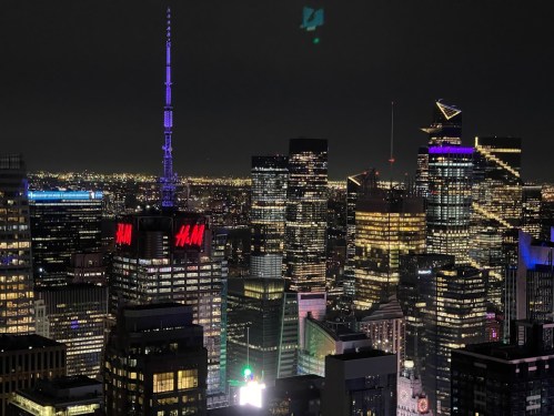A nighttime cityscape of skyscrapers, featuring illuminated buildings and a prominent H&M sign.