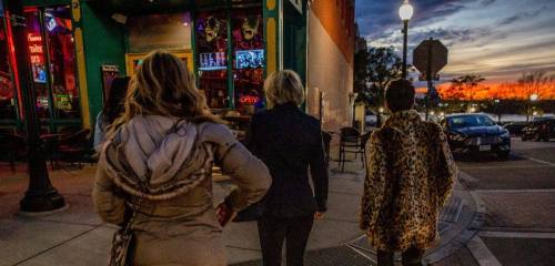 Three women walk down a city street at sunset, with colorful lights from nearby shops illuminating the scene.