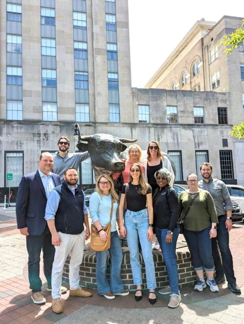 A group of people poses together in front of a bull statue in an urban setting on a sunny day.