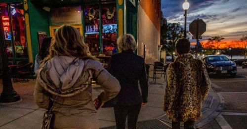 Three women walk down a city street at sunset, with colorful lights from nearby shops illuminating the scene.