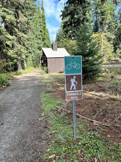 A trail sign indicating the Peninsula Trail with a bike symbol, surrounded by trees and a cabin in the background.