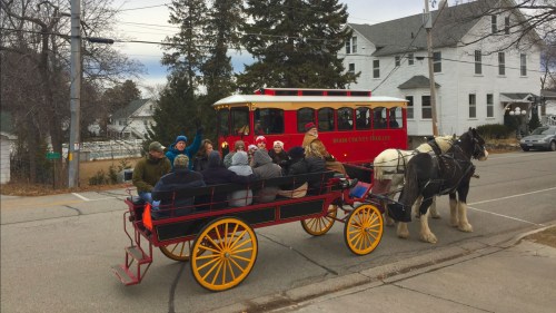 A horse-drawn carriage with passengers rides alongside a vintage trolley in a quaint town setting.