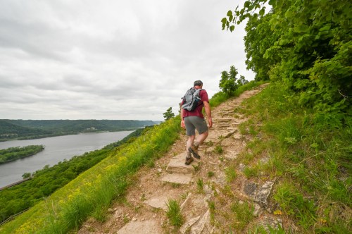 A hiker ascends a rocky trail beside a river, surrounded by lush greenery and overcast skies.