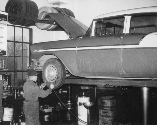 A mechanic works on a classic car lifted on a hydraulic rack in a vintage garage, surrounded by tires and tools.