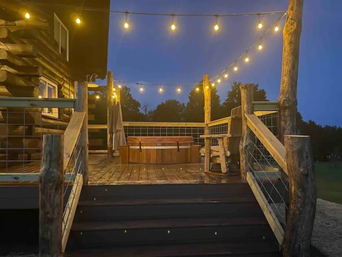 A cozy wooden deck at night, illuminated by string lights, featuring a hot tub and rustic seating.