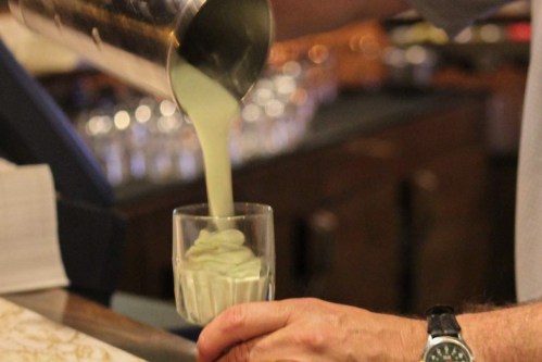 A person pours a creamy green beverage into a glass, with blurred glasses in the background.