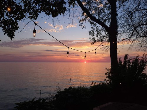 A serene sunset over a calm lake, with string lights hanging from a tree and colorful clouds in the sky.