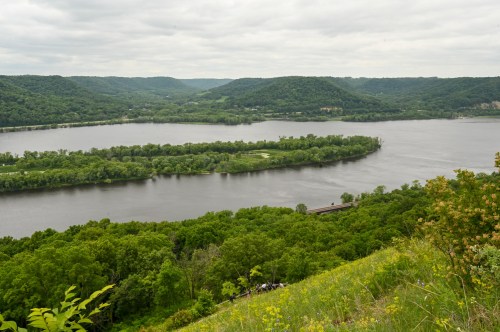 A scenic view of a winding river surrounded by lush green hills and trees under a cloudy sky.