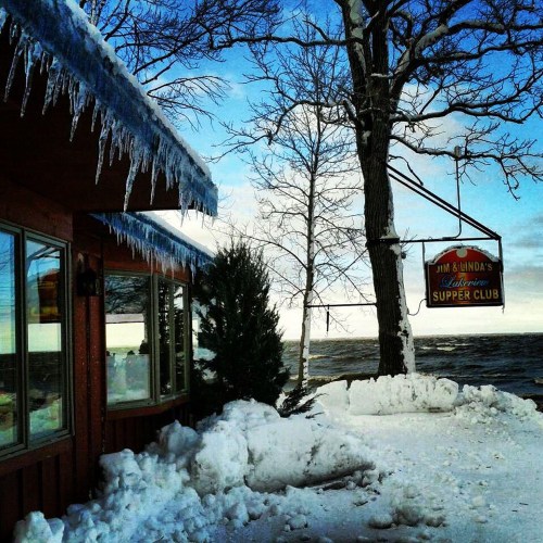 A cozy supper club by a snowy lake, with icicles hanging from the roof and a sign swaying in the winter breeze.