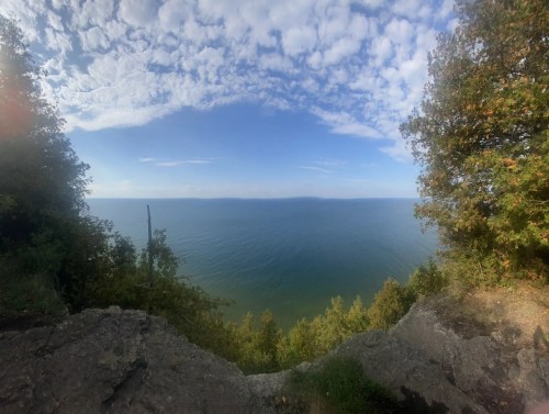 A panoramic view of a calm lake under a partly cloudy sky, framed by trees and rocky terrain.
