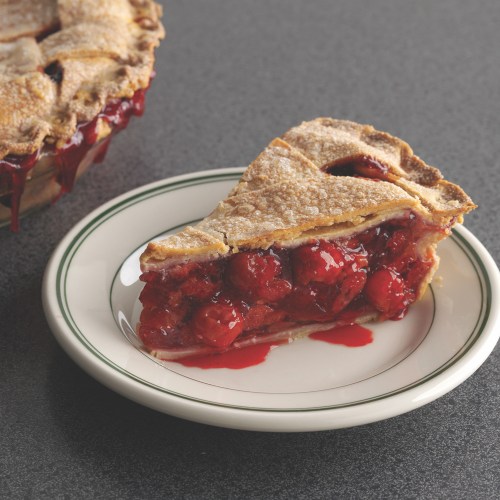 A slice of cherry pie on a plate, with a whole pie in the background, oozing with red cherry filling.