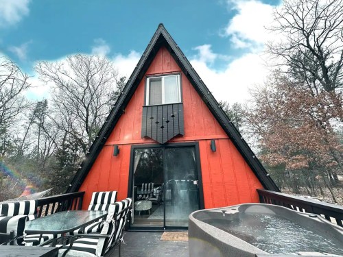 A red A-frame cabin with a hot tub on the deck, surrounded by trees under a blue sky with clouds.