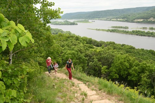 Three hikers ascend a rocky path surrounded by lush greenery, overlooking a river and valley below.