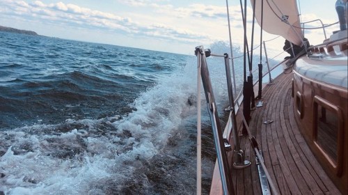 A sailboat glides through choppy waters, with waves splashing against the side and a cloudy sky above.