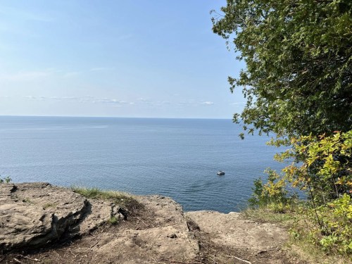 A serene view of a calm lake from a rocky cliff, with a small boat in the distance and trees framing the scene.