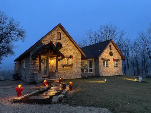 A cozy stone house decorated with Christmas wreaths and lights, surrounded by trees in a misty evening setting.