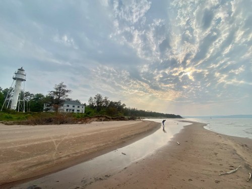 A sandy beach with a lighthouse in the background, a person walking along the shore under a cloudy sky.
