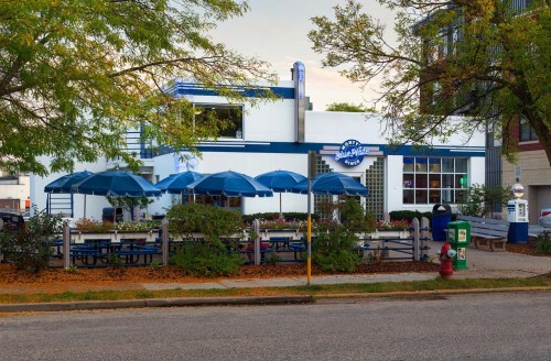 A retro-style diner with blue umbrellas and outdoor seating, surrounded by trees and autumn leaves.