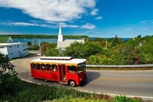 A red trolley bus drives along a scenic road with greenery and a church in the background, overlooking a body of water.