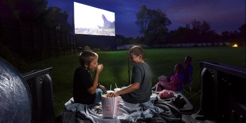 Two boys sit in the back of a truck, enjoying popcorn and drinks while watching a movie on a large outdoor screen.