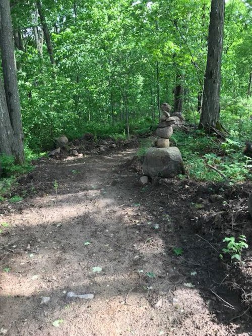 A dirt path through a lush green forest, featuring a stacked stone sculpture on the side.