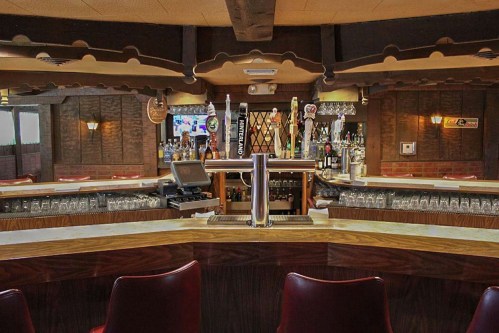 A cozy bar interior featuring a wooden counter, shelves of liquor, and red chairs in a warmly lit setting.