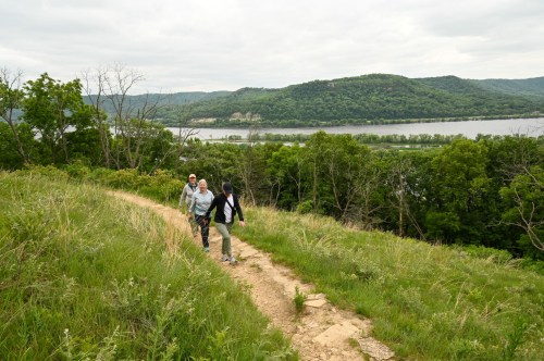 Three people walk along a grassy path with a river and hills in the background under a cloudy sky.