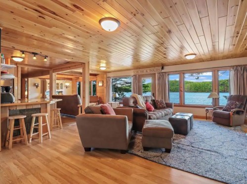 Cozy wooden living room with large windows, comfortable seating, and a view of the water. Warm, inviting atmosphere.