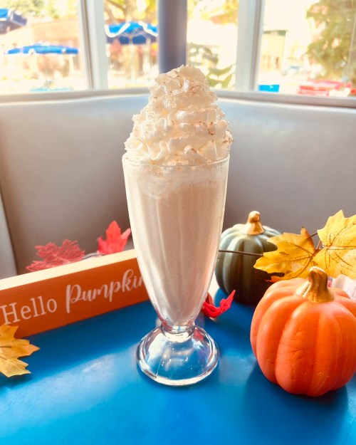 A tall glass of pumpkin-flavored milkshake topped with whipped cream, surrounded by autumn decorations.