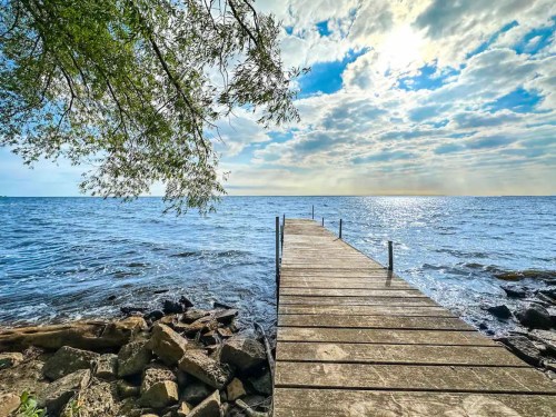 A wooden dock extends into a calm lake under a partly cloudy sky, surrounded by trees and rocky shoreline.