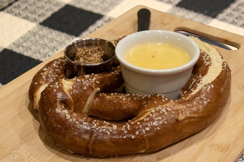 A large pretzel served on a wooden board with a small bowl of cheese dip and a side of mustard.