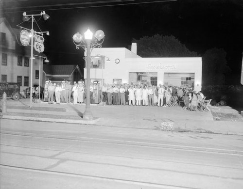 A crowd gathers outside a vintage service station at night, with classic cars parked nearby and streetlights illuminating the scene.