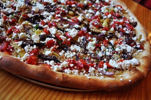 A close-up of a pizza topped with vegetables, herbs, and crumbled cheese on a wooden table.