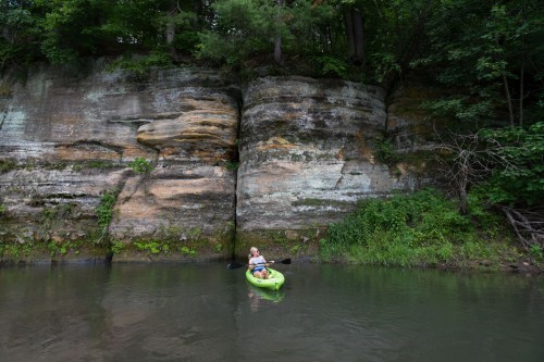 A person in a kayak floats on calm water near a rocky cliff surrounded by lush greenery.