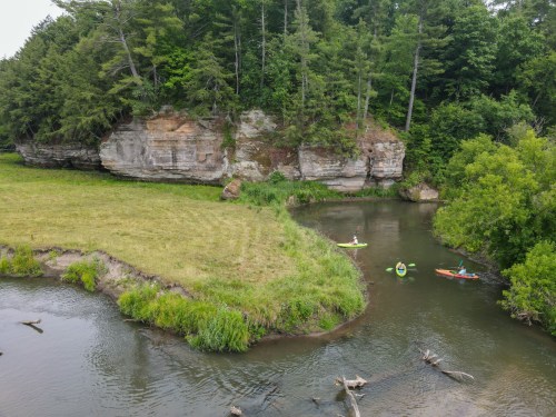 Two kayakers paddle on a winding river surrounded by lush greenery and rocky cliffs.