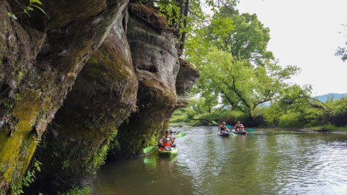 Two kayakers paddle along a serene river, flanked by lush greenery and a large rock formation.