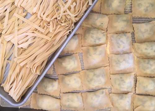 A tray of freshly made pasta, including long noodles and square ravioli, arranged neatly on a surface.