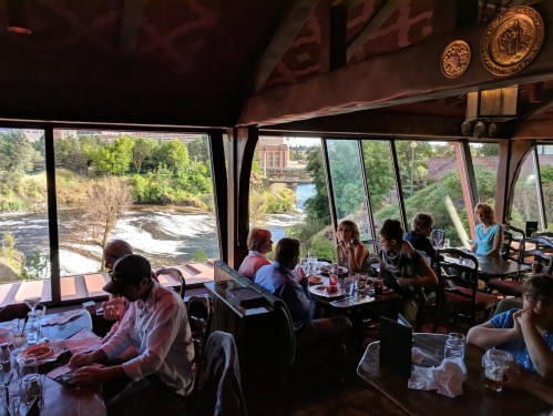 A restaurant interior with diners enjoying meals, large windows overlooking a river and greenery outside.