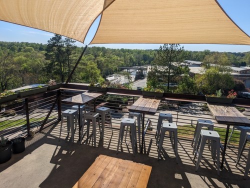 A rooftop patio with metal stools and wooden tables, overlooking a green landscape and a distant road.