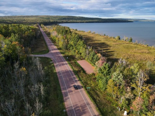 Aerial view of a winding road alongside a lake, surrounded by trees and grassy areas under a partly cloudy sky.