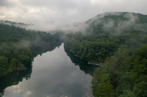 A serene river surrounded by lush green trees and misty mountains, reflecting the cloudy sky above.