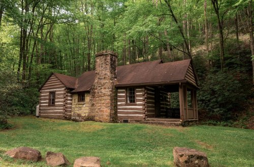 A rustic log cabin with a stone chimney, surrounded by lush green trees and grass.