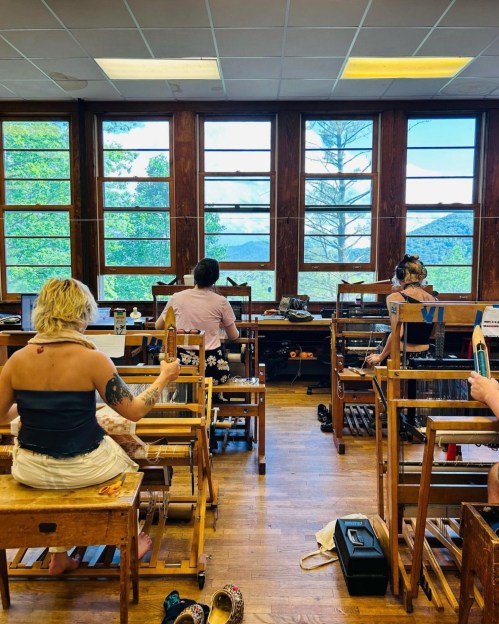 A group of people working at wooden looms in a bright room with large windows overlooking greenery.