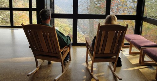 Two people sit in rocking chairs by a large window, overlooking a scenic view of mountains and autumn foliage.