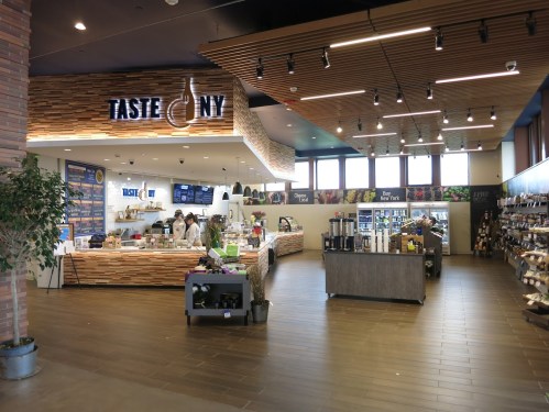 Interior of a modern café named "Taste NY," featuring a counter, seating area, and shelves with various products.