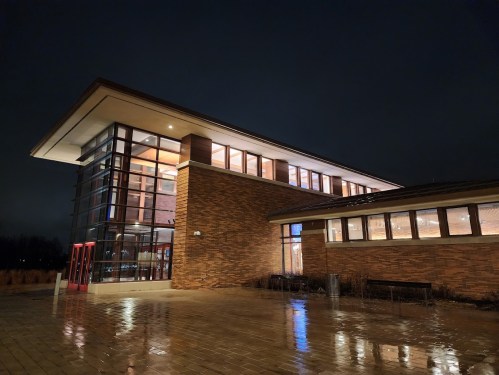 Modern brick building illuminated at night, featuring large windows and a sleek design, with wet pavement reflecting light.